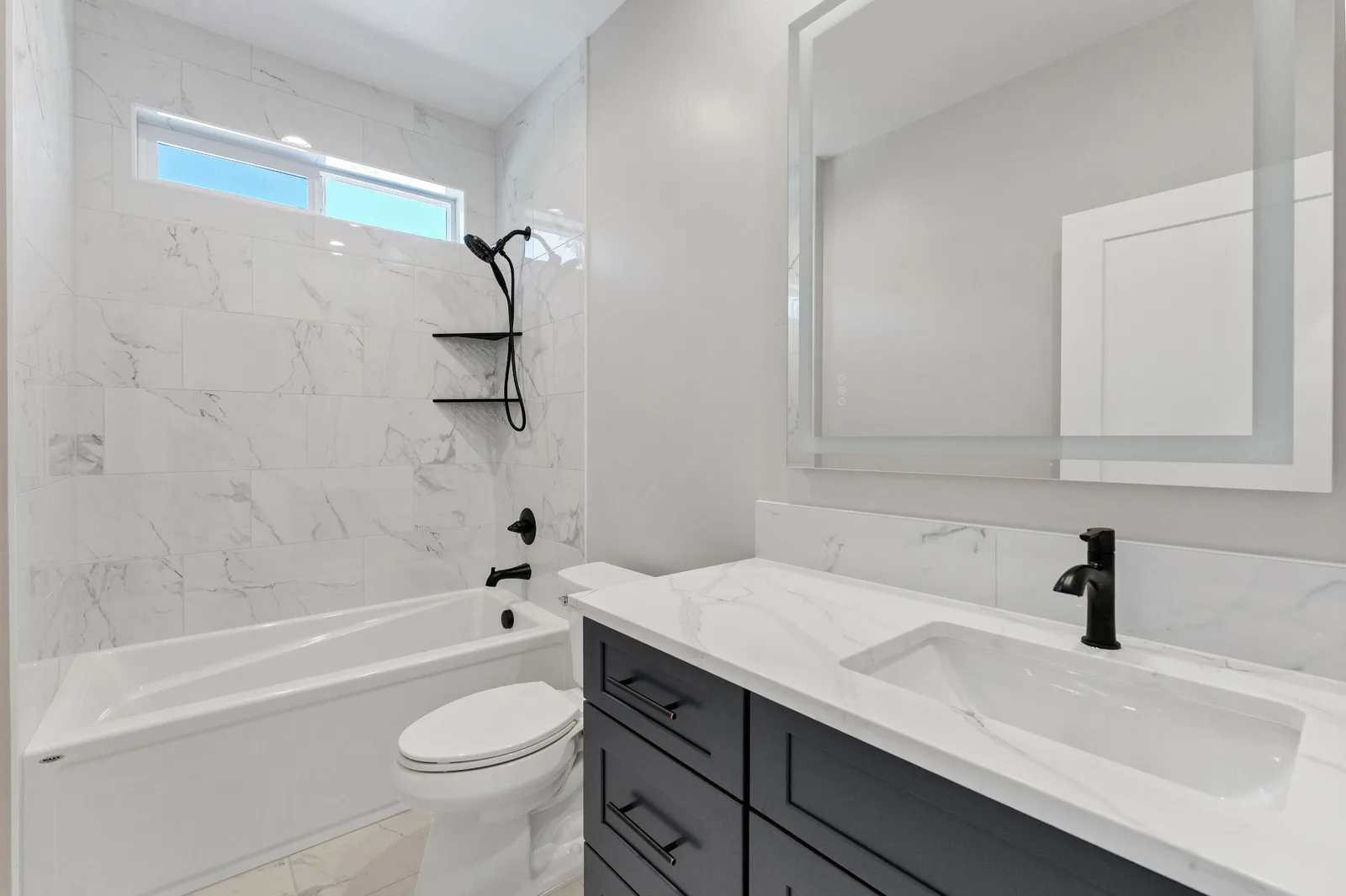 Guest bath with marble-look tile surround, navy vanity and lit mirror