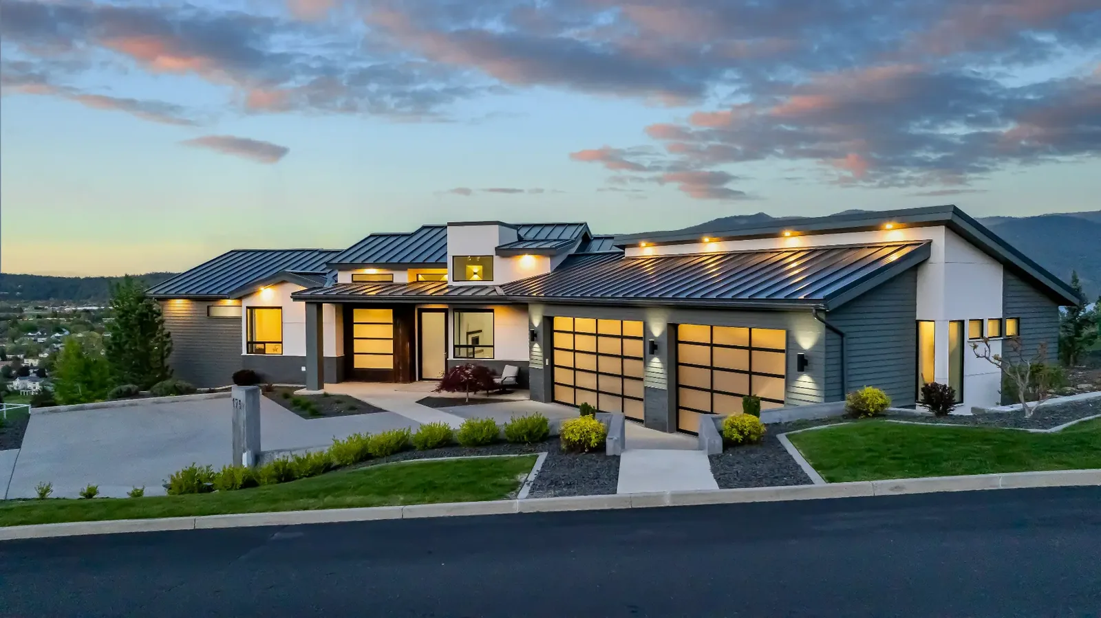 Front elevation at twilight with standing-seam roof and lit glass garage doors